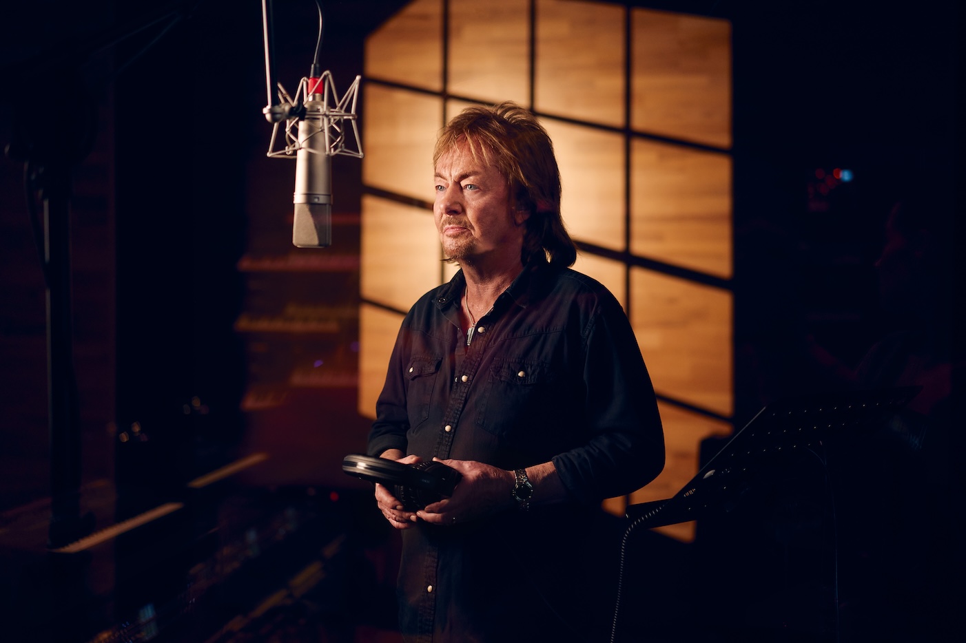Chris Norman holds headphones in his hands and stands in front of a microphone.